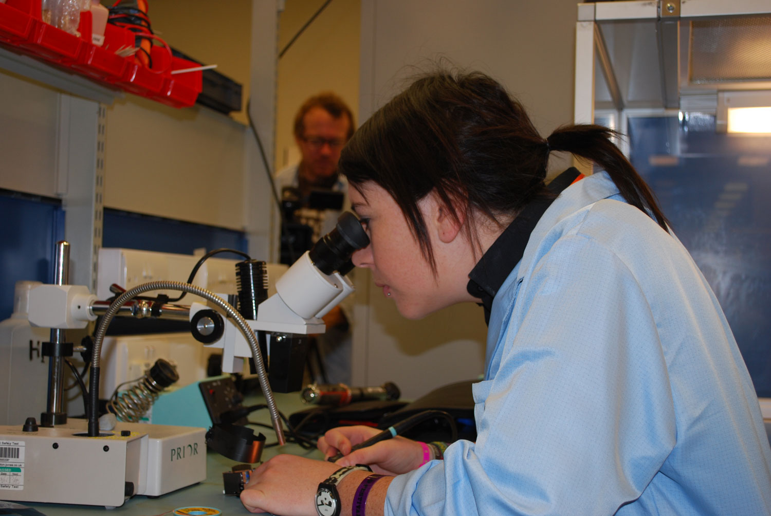 A person with dark brown hair in a lab coat leaning over and looking into a microscope.