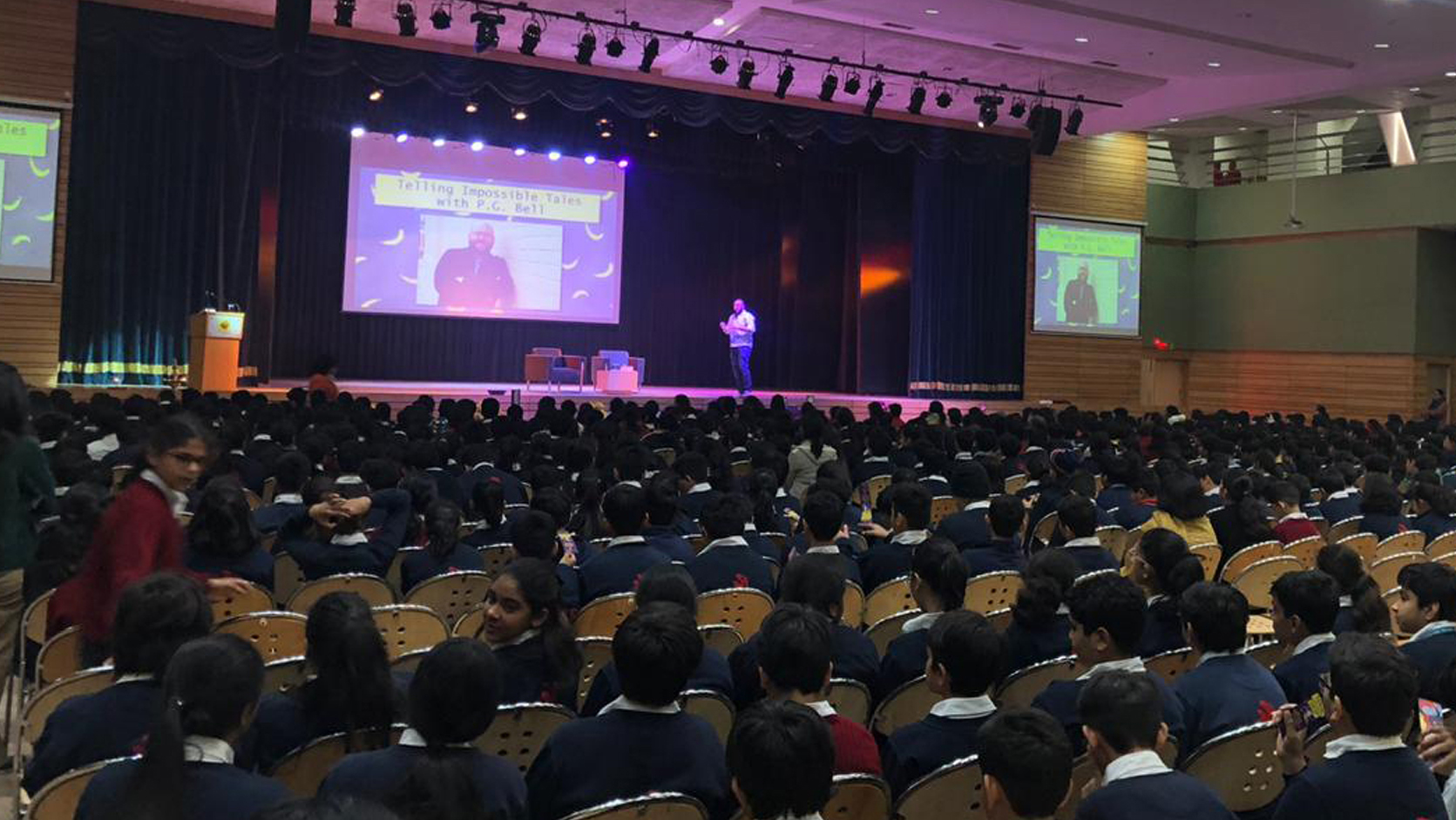 A group of school children sitting down in a large hall watching a large screen on a stage.