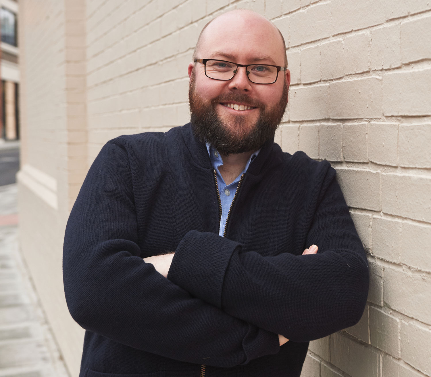 A man leaning against a brick wall wearing a blue shirt and navy jumper smiling.